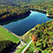 An aerial image of a large still blue lake surrounded by a dense forest of autumnal trees