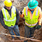 Two people in safety vests and hard hats standing in a deep muddy hole discussing visible exposed cables