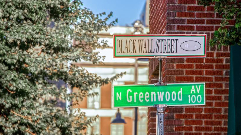 Street signs for Black Wall Street and Greenwood Avenue near a brick building