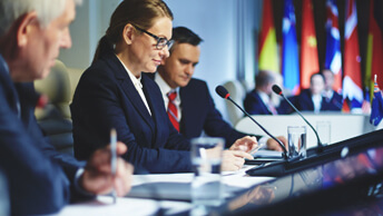 People sitting next to each other at a table with microphones and flags in the background
