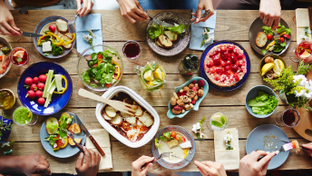 Many colorful plates of food on a long wood table surrounded by the hands of the people sitting at the table