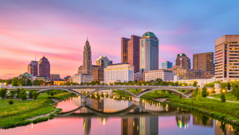 A city skyline towers over a bridge and reflective river at twilight