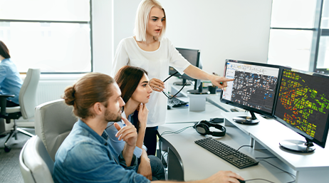 Three business professionals pointing to and looking at maps on computer monitors