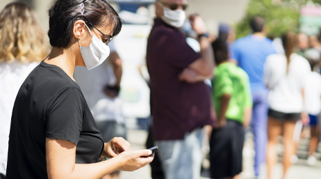 A person wearing a face mask that covers their nose and mouth looking at their phone while standing in a line of people