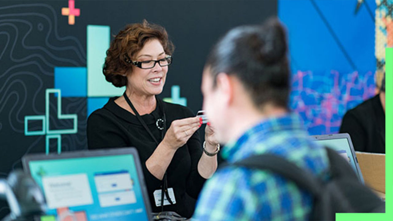 A photo of a conference staffer helping a registrant, while a person in a backpack stands in the foreground using a laptop